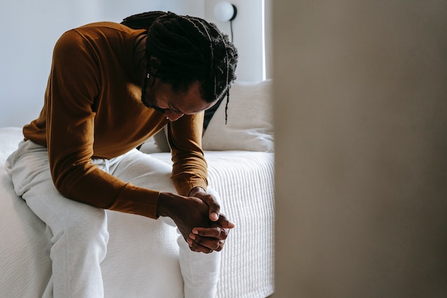 man sitting on bed with hands folded on his lap as he stares down at floor